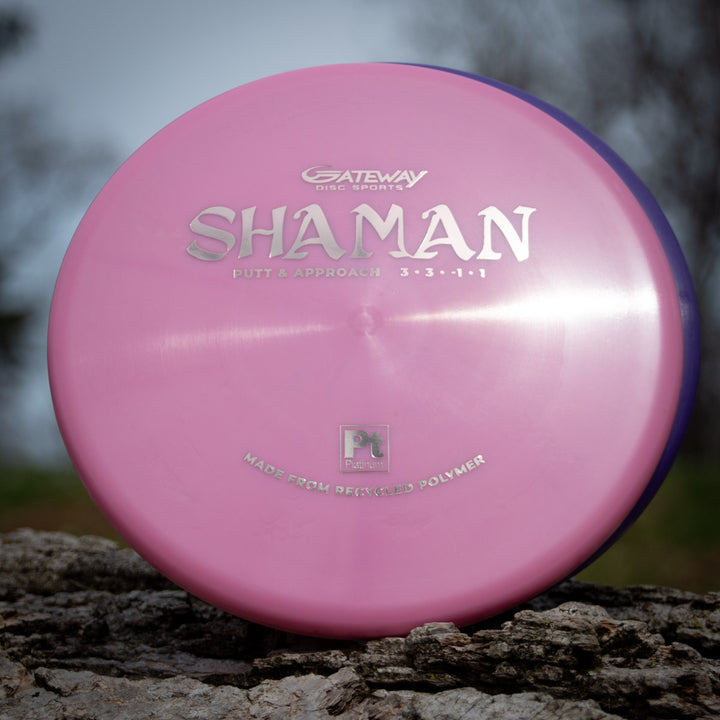 A pink Shaman - Platinum disc golf putter made from recycled plastic rests on a tree stump, its silver text highlighting its sustainable design.