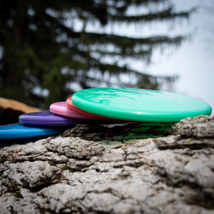 A close-up of four Shaman - Platinum disc golf discs made from recycled plastic, stacked on a tree trunk with blurred trees in the background—a sustainable choice for eco-conscious players.