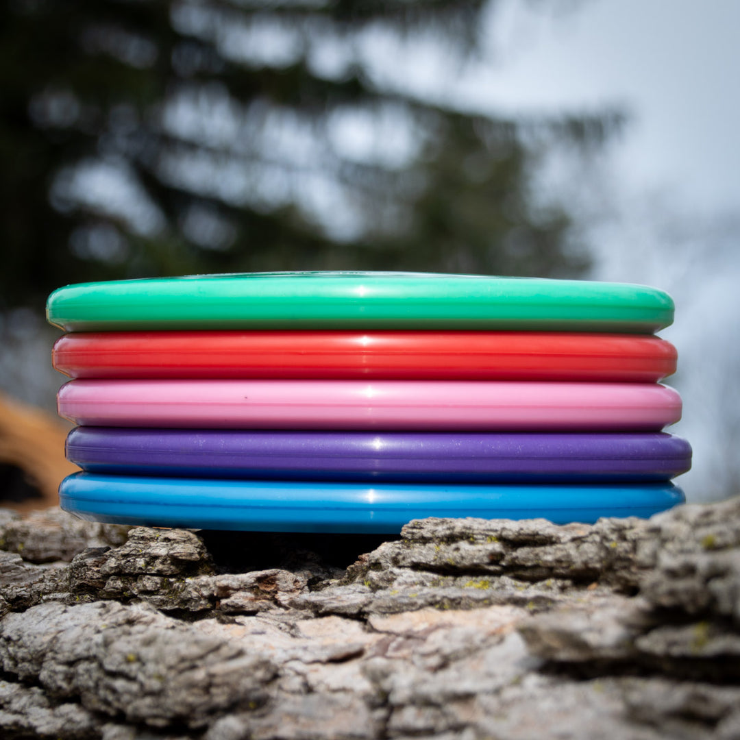 A stack of five vibrant Shaman - Platinum discs—green, red, pink, purple, and blue—crafted from recycled plastic rests on a rugged tree trunk outdoors with blurred trees behind.