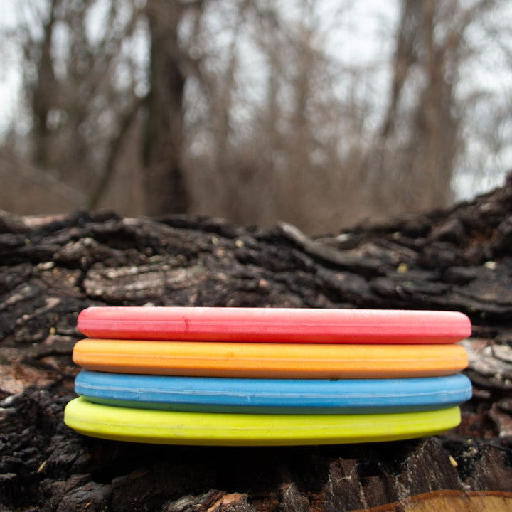 Four brightly colored disc golf discs—including a Shaman - Suregrip with no bead and a 25° flat nose—are stacked on a log, with blurred trees in the background.