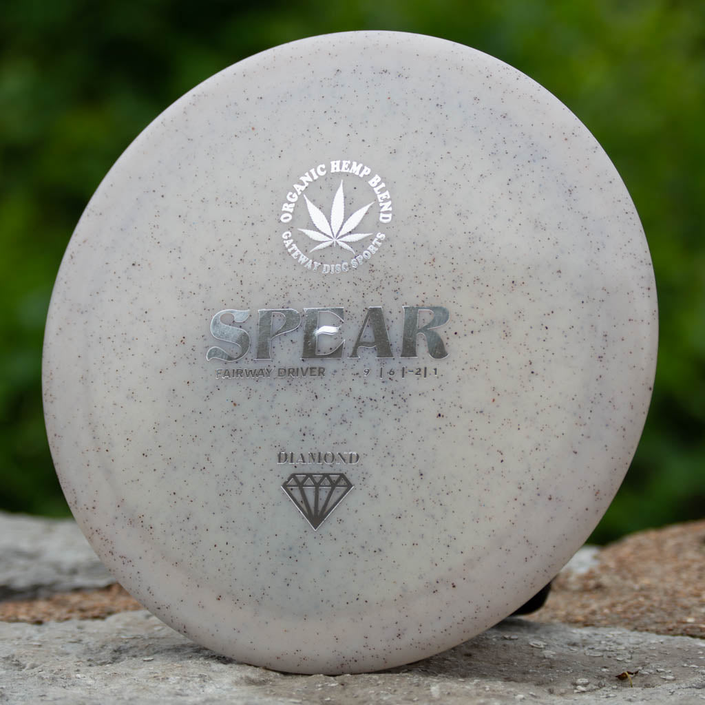 The Spear - Diamond Hemp control driver features a white speckled look with the organic hemp blend logo and diamond symbol, resting on stone among greenery—perfect for accurate throws on the course.