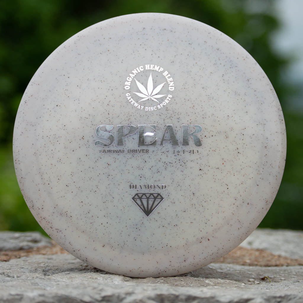A white Spear - Diamond Hemp control driver disc golf disc featuring —SPEAR,—� —Organic Hemp Blend,—� and a diamond icon, rests on stone with greenery in the background—designed for accurate placement.