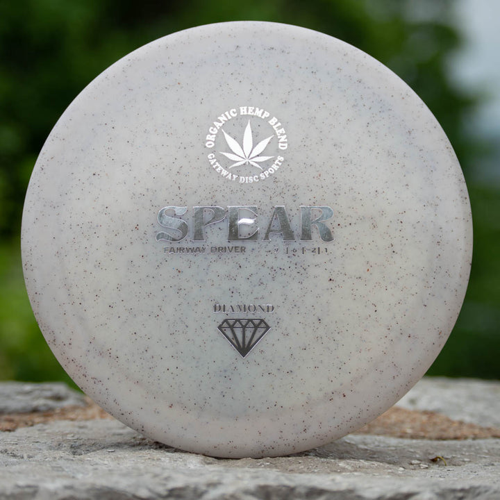 A white Spear - Diamond Hemp control driver disc golf disc featuring —SPEAR,—� —Organic Hemp Blend,—� and a diamond icon, rests on stone with greenery in the background—designed for accurate placement.