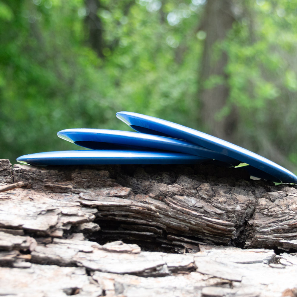 Three Spear - Nylon blue discs, perfect as control drivers, are stacked on rough tree bark with their nylon blends shining softly against a blurred green forest background.
