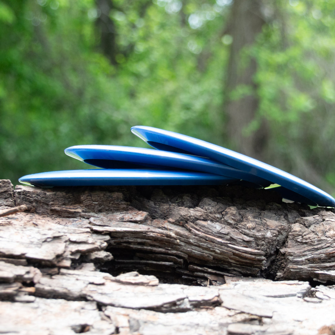 Three Spear - Nylon blue discs, perfect as control drivers, are stacked on rough tree bark with their nylon blends shining softly against a blurred green forest background.