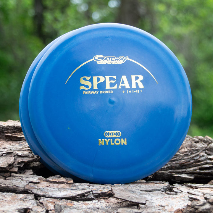 A blue Spear - Nylon control driver disc by Gateway Disc Sports, made with nylon blends and labeled "NYLON," rests on tree bark, promising a straight flight against a blurred green background.