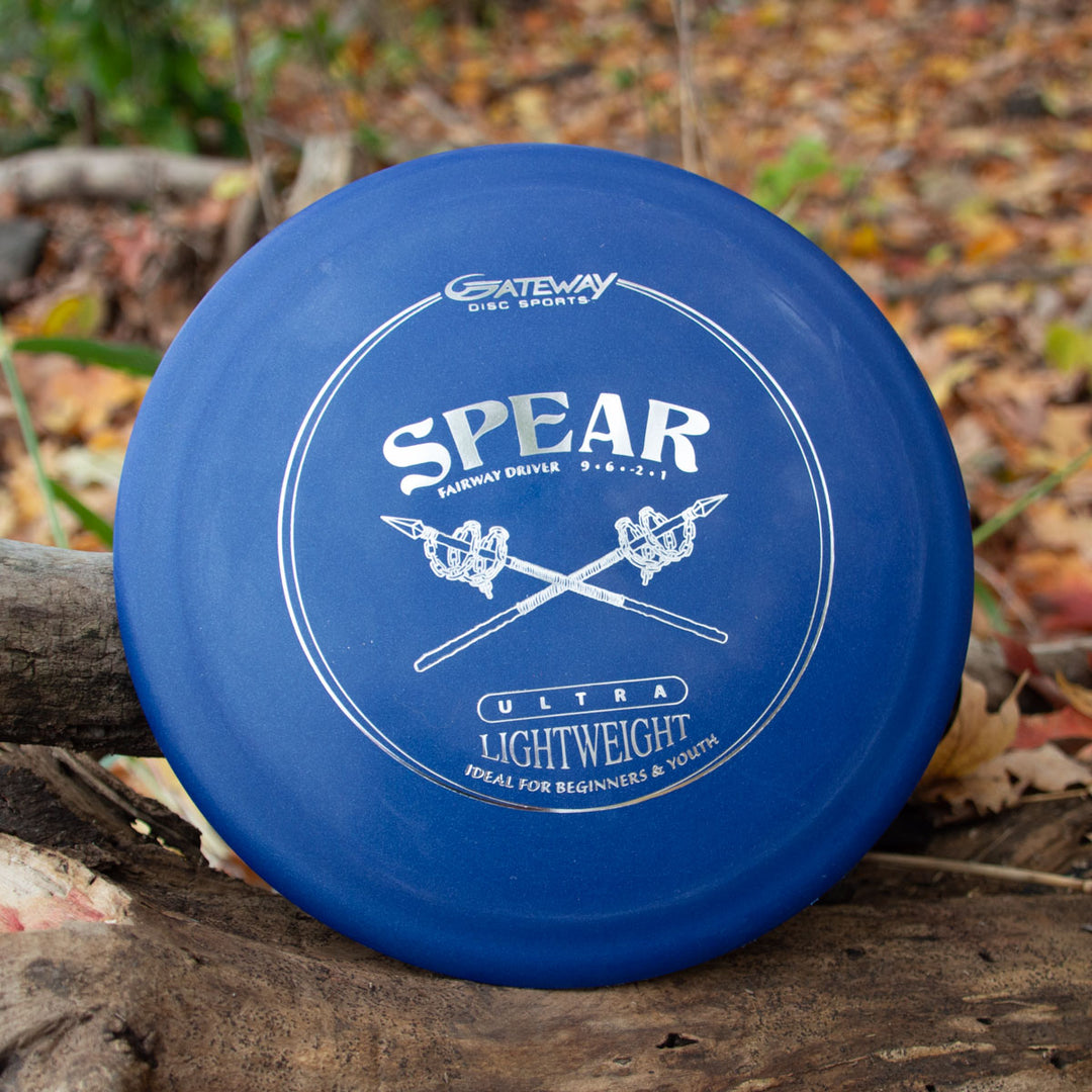 A blue Spear - SureGrip™  Ultra Lightweight control driver disc by Gateway Disc Sports rests on a log among fallen leaves, ready for precise placement on the course.