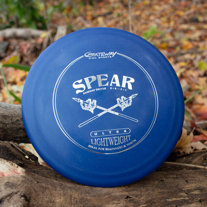A blue Spear - SureGrip™  Ultra Lightweight control driver disc by Gateway Disc Sports rests on a log among fallen leaves, ready for precise placement on the course.