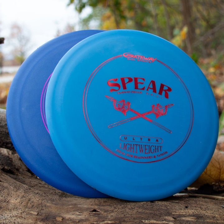 Two blue disc golf discs rest on a log outdoors; one is labeled "Spear - SureGrip™  Ultra Lightweight" in red text and graphics—a control driver made for precise throws. Trees and foliage appear in the background.