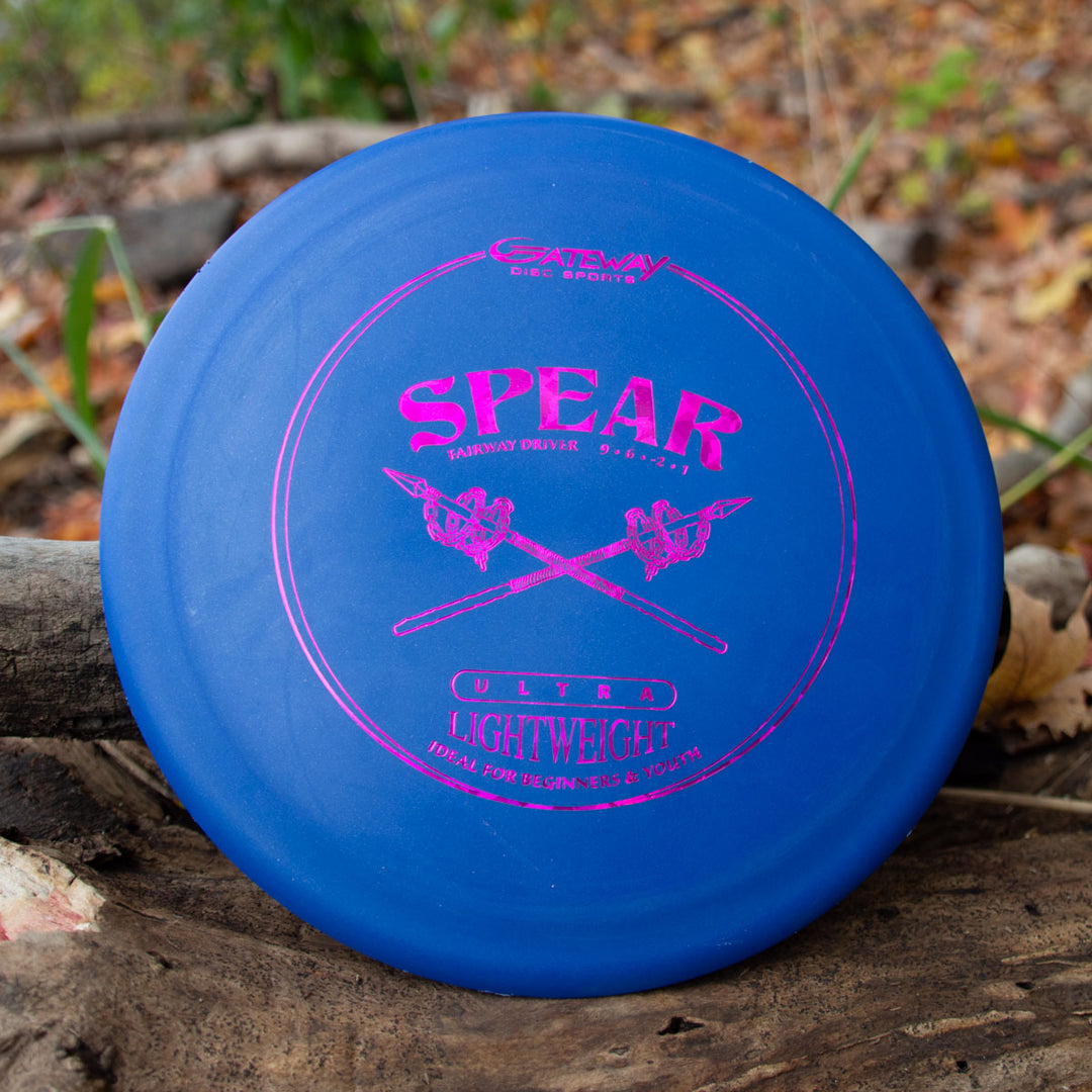A blue Spear - SureGrip™  Ultra Lightweight control driver disc with pink text and graphics sits on a log in the woods, perfect for accurate placement on the course.