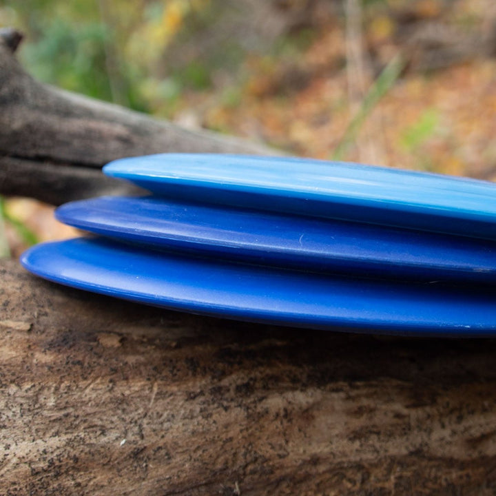 Three blue Spear - SureGrip™  Ultra Lightweight driver discs stacked on a log outdoors, set against a blurred natural background—ideal for players seeking accuracy and maximum distance.