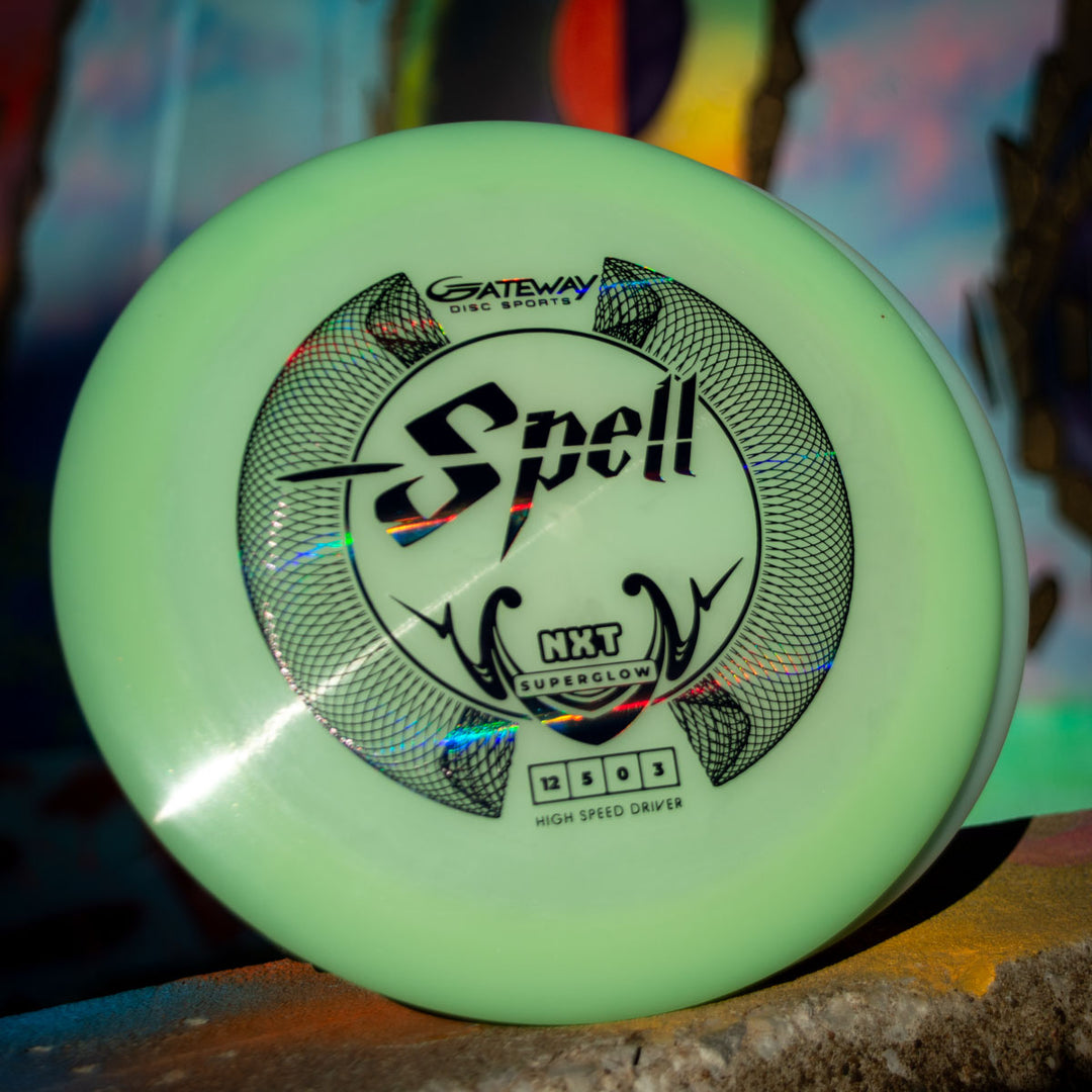 A Spell - NXT Superglow overstable driver from Gateway Disc Sports is shown upright on a surface with a decorative pattern, set against a vibrant, blurred background, highlighting its wind-crushing power and superior durability.