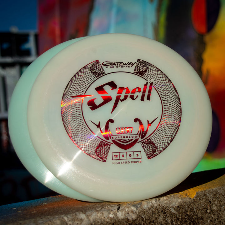 Close-up of a white Spell - NXT Superglow overstable driver by Gateway Disc Sports with red graphics, set against a vibrant, blurred background, highlighting its wind-crushing performance.