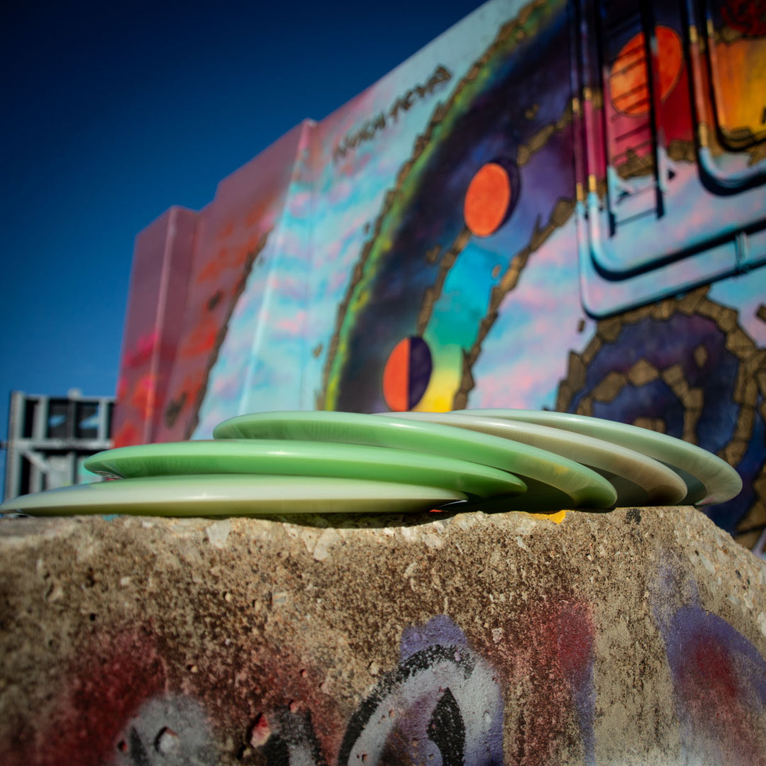 Four Spell - NXT Superglow green and white disc golf discs, including a durable overstable driver, rest on concrete in front of a colorful graffiti mural with abstract patterns and circles.