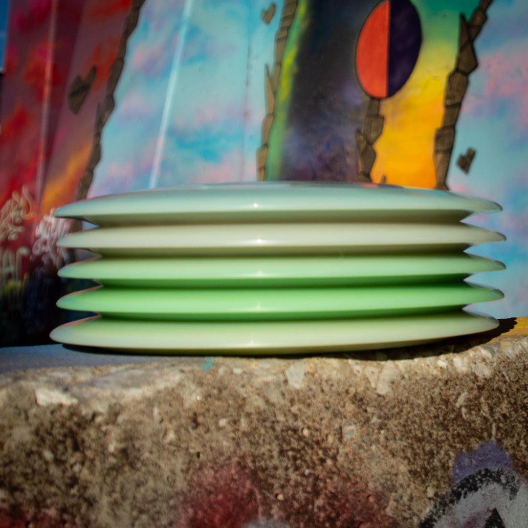 Six Spell - NXT Superglow pale green disc golf discs, prized for their durability, are stacked on a concrete ledge with colorful graffiti in the background.