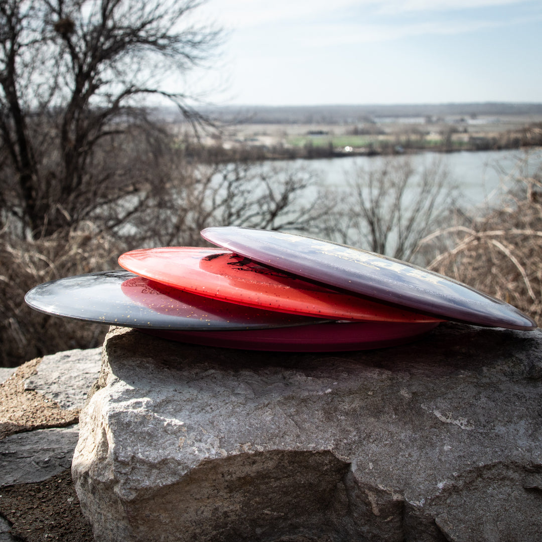 Three Gateway Spell - Metal Flake - Ripper Studios discs—two red and one black—crafted from premium plastic, are stacked on a large stone with a river and leafless trees in the background.