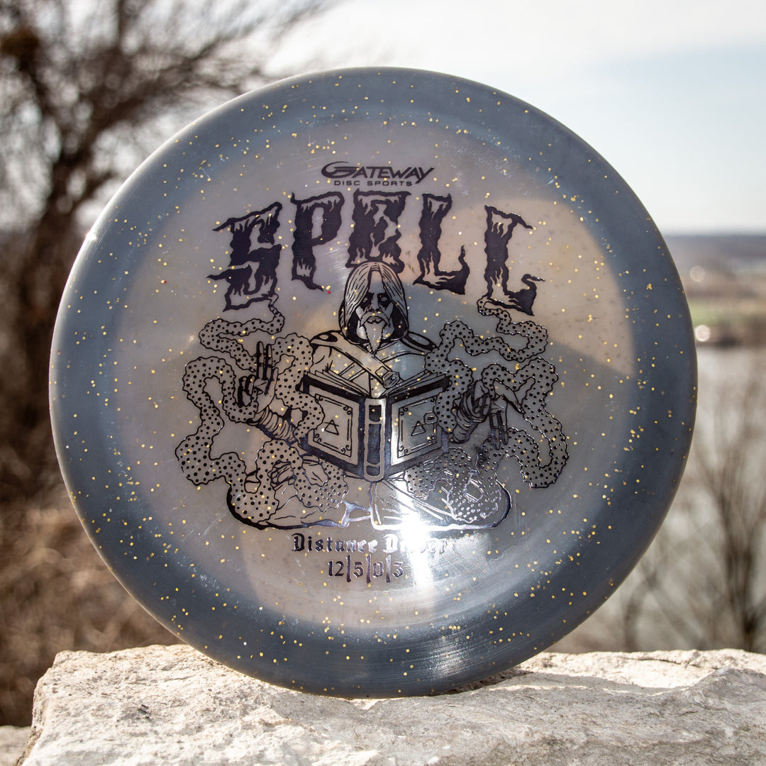 The Spell - Metal Flake - Ripper Studios disc by Gateway Disc Sports, in gray and gold speckle, features a wizard, open book, and snakes—crafted from premium plastic for legendary durability and distance.