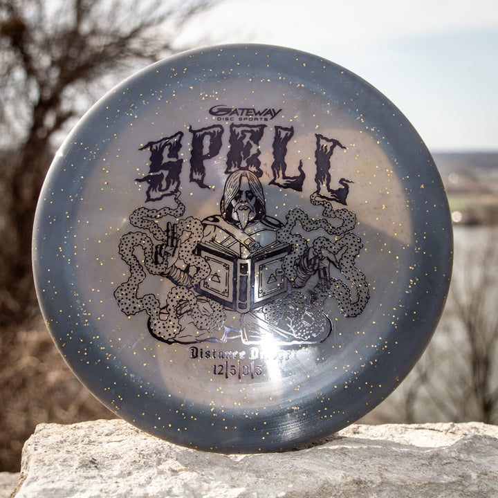 The Spell - Metal Flake - Ripper Studios disc by Gateway Disc Sports, in gray and gold speckle, features a wizard, open book, and snakes—crafted from premium plastic for legendary durability and distance.