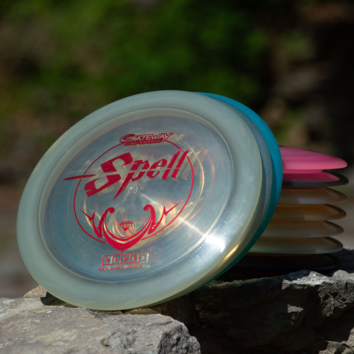A stack of disc golf discs, featuring the overstable "Spell - Diamond" driver with red text, sits on a rock outdoors—ideal for powerful, wind-crushing throws.