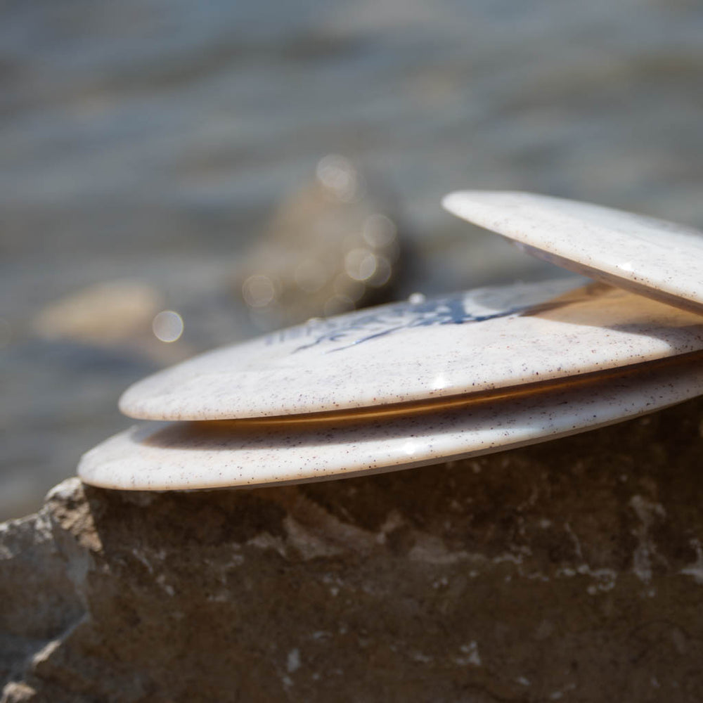 Three Spell - Diamond Hemp white disc golf discs rest on a large rock by the water's edge, ready for a high-velocity throw, with blurred stones in the background.