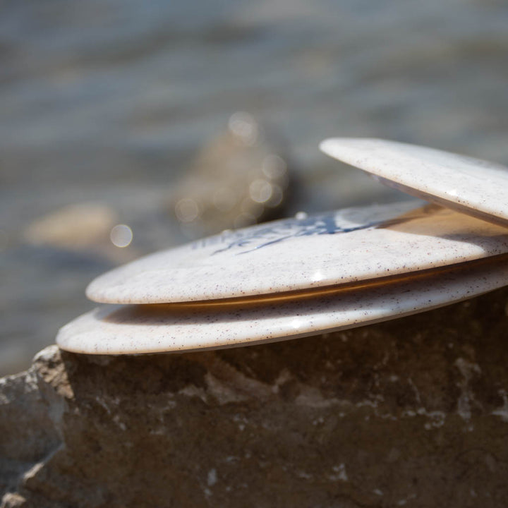 Three Spell - Diamond Hemp white disc golf discs rest on a large rock by the water's edge, ready for a high-velocity throw, with blurred stones in the background.