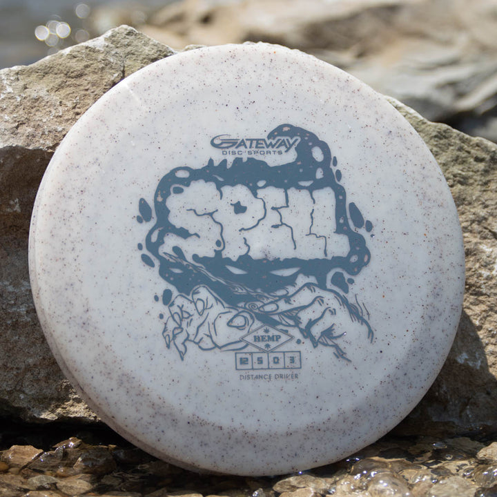 A white Gateway "Spell - Diamond Hemp" distance disc, made from premium hemp plastic with gray graphics, lies on rocks by the water—poised for a powerful throw.