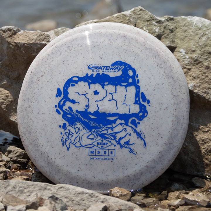 A white speckled Spell - Diamond Hemp flying disc golf disc with blue artwork and text, made from premium plastic, rests against rocks near water.