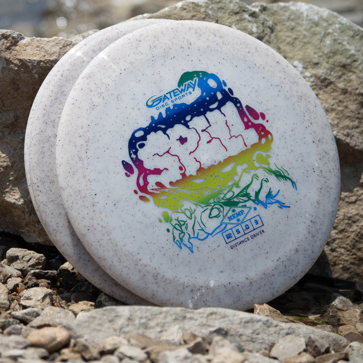 A white Spell - Diamond Hemp disc golf driver with vibrant graphics, made from premium plastic, rests against a rock on small stones.