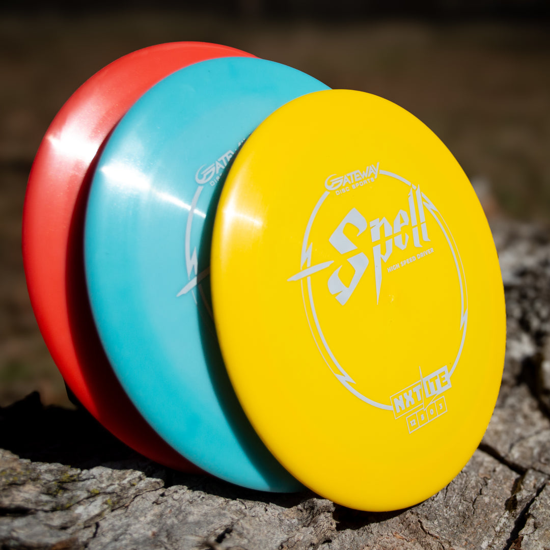 Three disc golf discs—red, blue, and the Spell - NXT Lite yellow overstable driver—stand stacked upright on a tree trunk outdoors. "Spell" and "Gateway" are printed on the yellow disc, ready to showcase wind-crushing power.