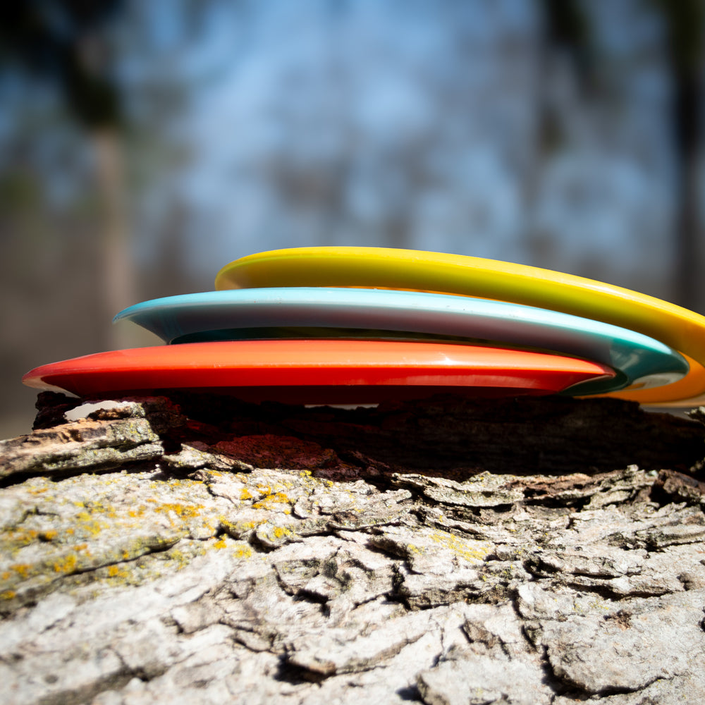 Four vibrant Spell - NXT Lite discs, including an overstable driver built for wind-crushing throws, are stacked on a tree trunk against a softly blurred outdoor backdrop.