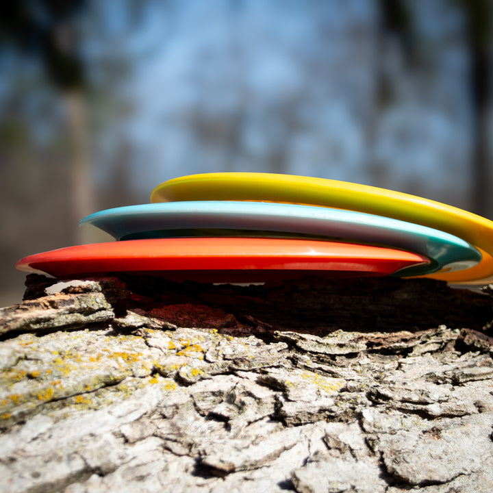 Four vibrant Spell - NXT Lite discs, including an overstable driver built for wind-crushing throws, are stacked on a tree trunk against a softly blurred outdoor backdrop.
