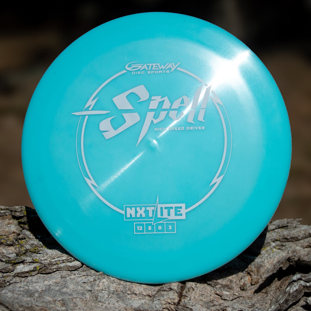 A turquoise Spell - NXT Lite overstable driver from Gateway Disc Sports rests on rough wood, its surface catching the sunlight and promising exceptional wind-fighting performance.