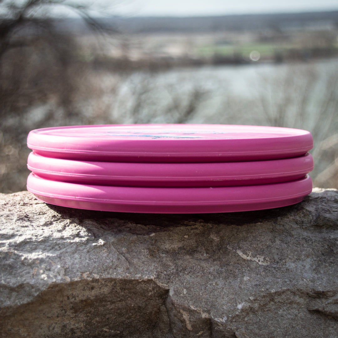 Three Reptilian DG Stego — Fossil pink discs, dubbed the infamous #WorstDiscEverMade, are stacked on a stone ledge against a blurred outdoor backdrop—an ideal shot to discuss their unique Flight Numbers.
