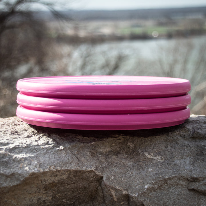 Three Reptilian DG Stego — Fossil pink discs, dubbed the infamous #WorstDiscEverMade, are stacked on a stone ledge against a blurred outdoor backdrop—an ideal shot to discuss their unique Flight Numbers.