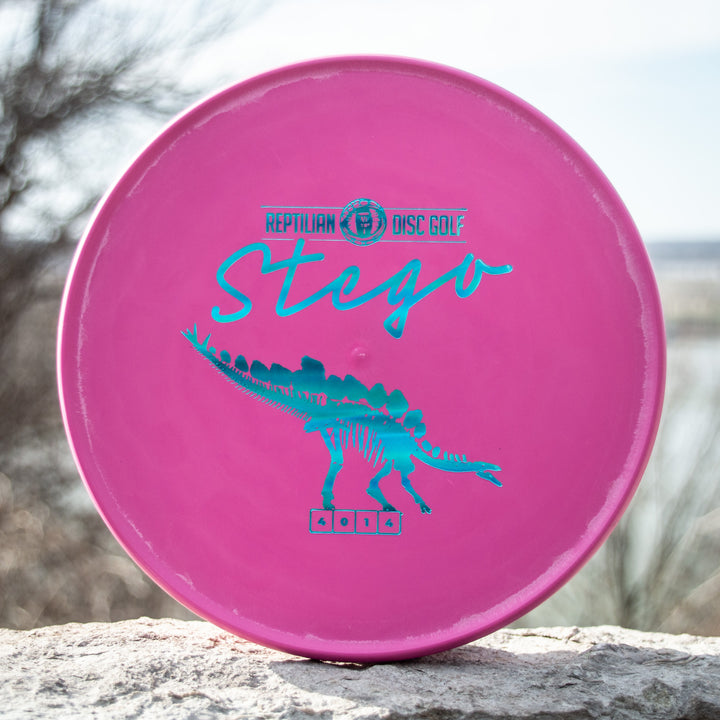 A pink Reptilian DG Stego — Fossil disc, featuring "Stego" and a teal stegosaurus graphic, lies on stone outdoors—often called the #WorstDiscEverMade for its unique Flight Numbers.