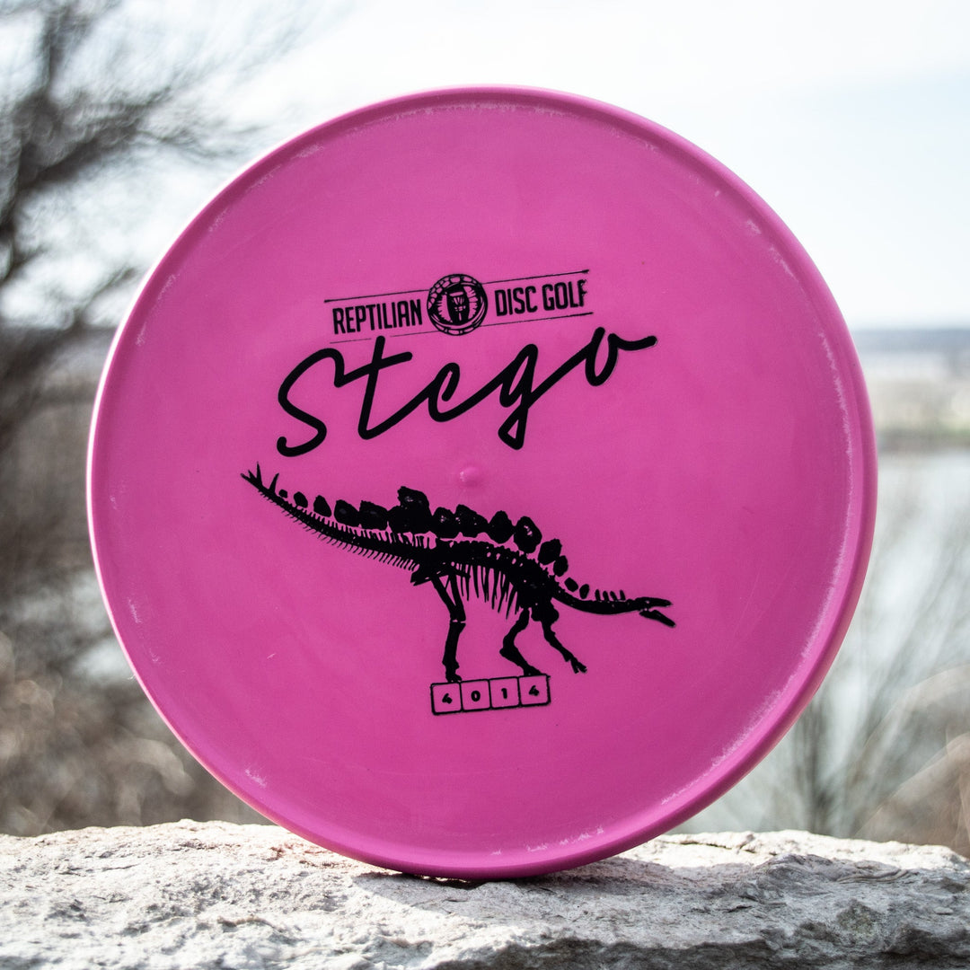 A pink Reptilian DG Stego — Fossil disc with its flight numbers and a black dinosaur skeleton graphic rests on stone outdoors—famously called by some the #WorstDiscEverMade.