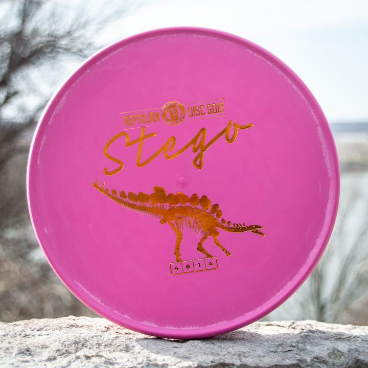 A pink Reptilian DG Stego — Fossil disc golf disc with a gold stegosaurus graphic and bold flight numbers 4 0 1 4, famously dubbed the #WorstDiscEverMade by some players.