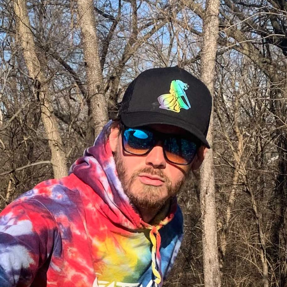 A man in a tie-dye hoodie, sunglasses, and the Rainbow Wizard SnapBack Hat with a front logo poses outside among leafless trees.