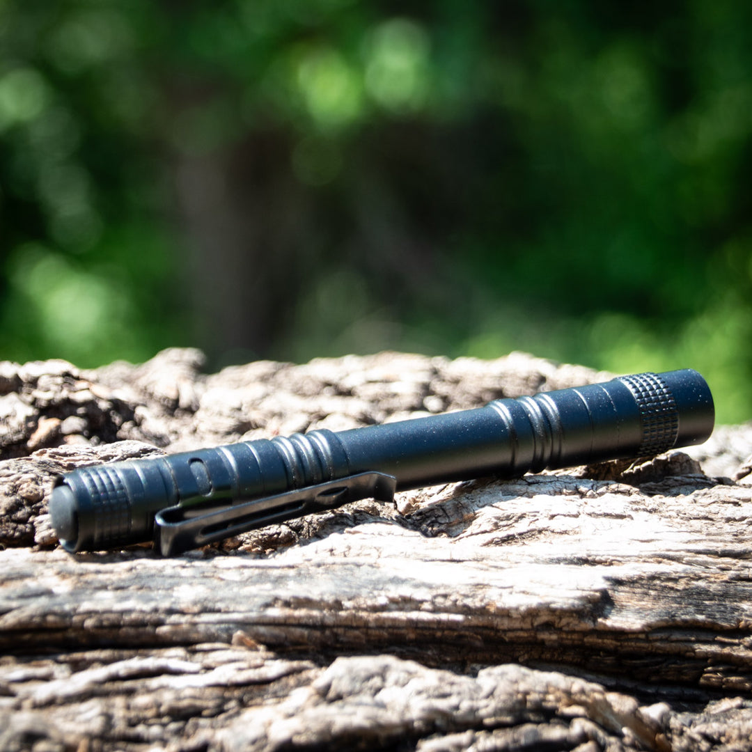 A UV Laser Pointer, shaped like a black pen with a clip, lies on rough textured wood while green foliage appears blurred in the background.
