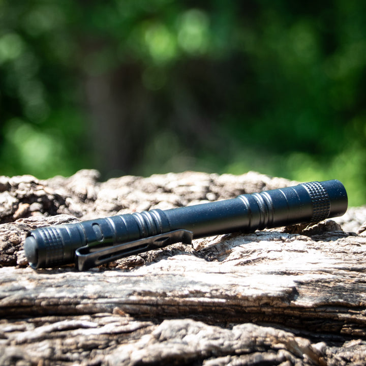 A UV Laser Pointer, shaped like a black pen with a clip, lies on rough textured wood while green foliage appears blurred in the background.