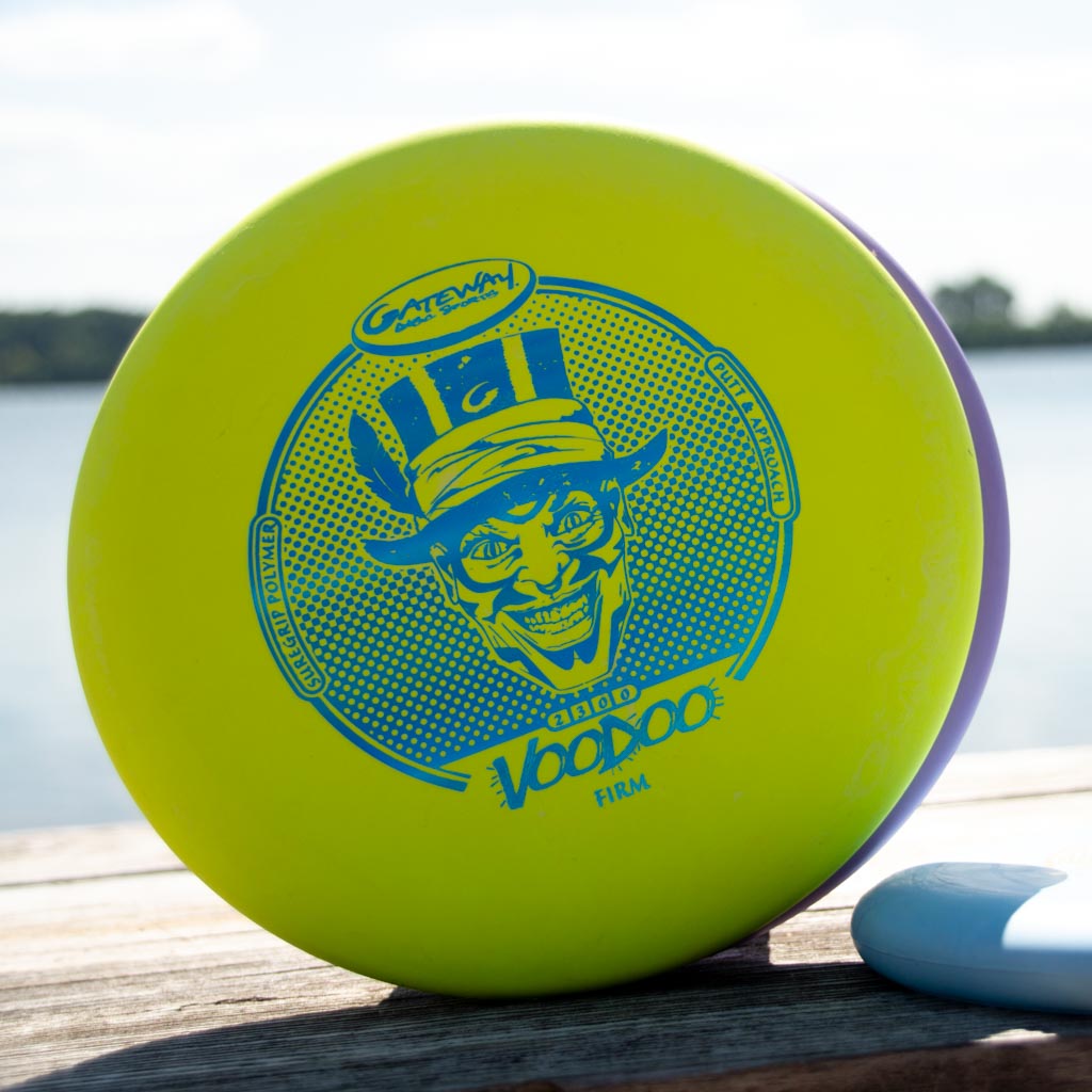 A yellow Voodoo - Suregrip - Firm disc golf putter with a blue grinning character graphic, displayed on wood beside water.