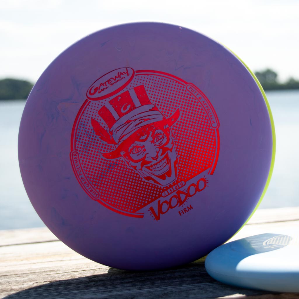 A Voodoo - Suregrip - Firm putter with a purple and red design sits on wood by the water, making it an eye-catching lay-up disc for your short game.