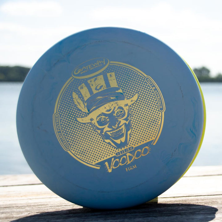A blue Voodoo - Suregrip - Firm putter with a gold skull-in-hat graphic rests on a wooden surface by the water—perfect for your next lay-up.