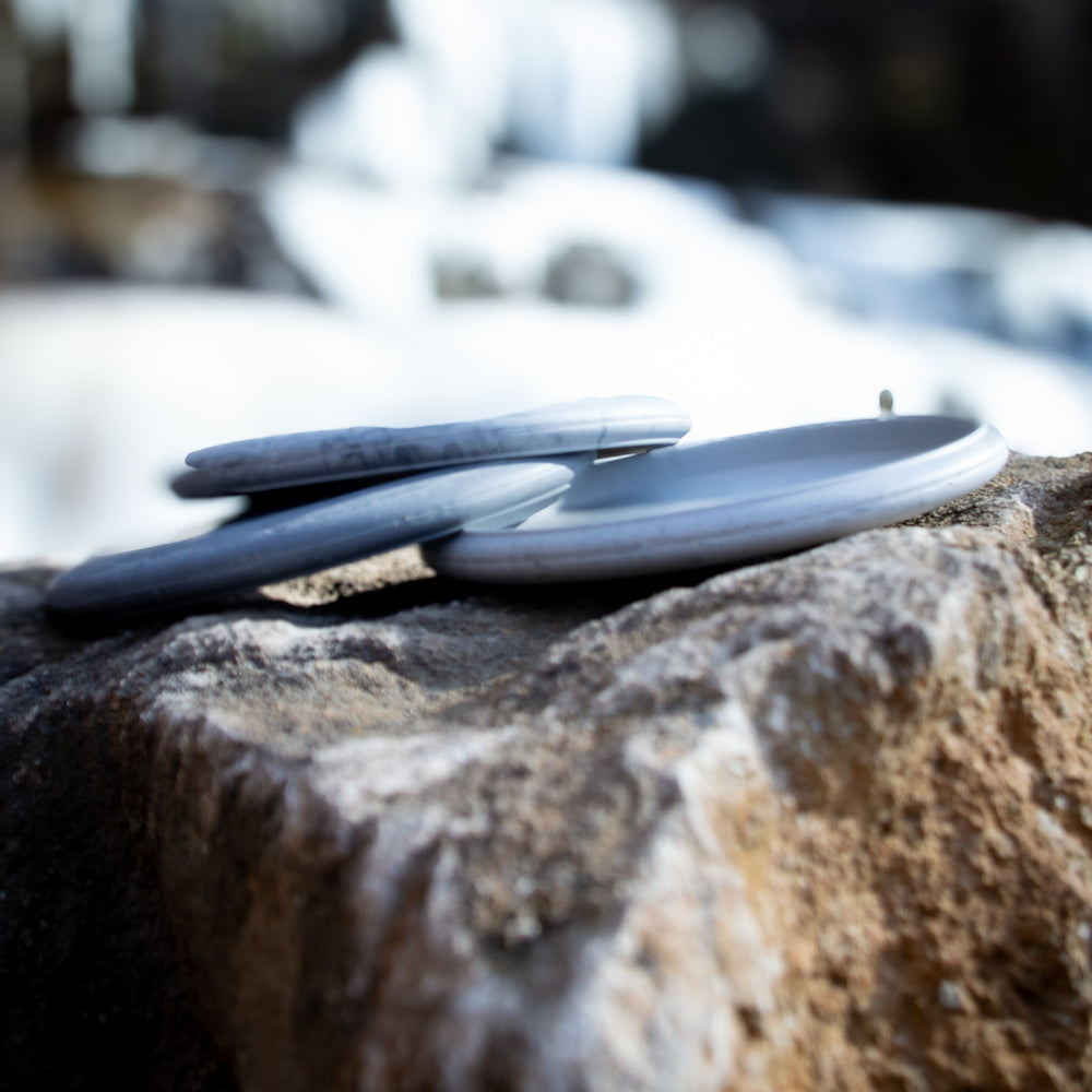 Several smooth, flat gray stones are stacked on a large rock outdoors, their disc-like shapes reminiscent of the Voodoo - Lunar putter, set against a softly blurred background.