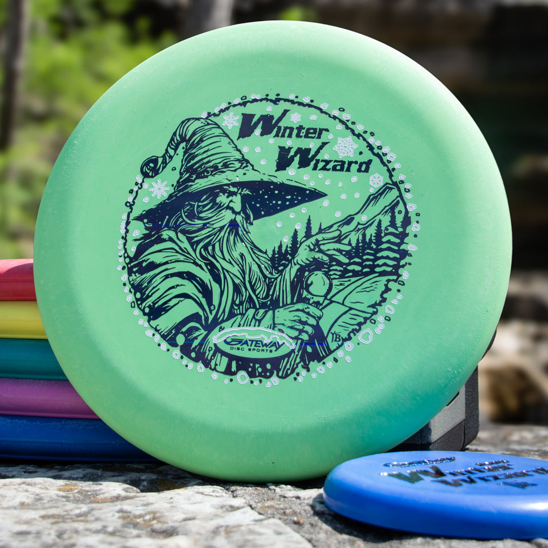 The Winter Wizard Wizard - SSSS + Mini is a green disc golf disc labeled "Winter Wizard" with a wizard illustration, accompanied by a mini—ideal for players seeking max weight and great flight performance.