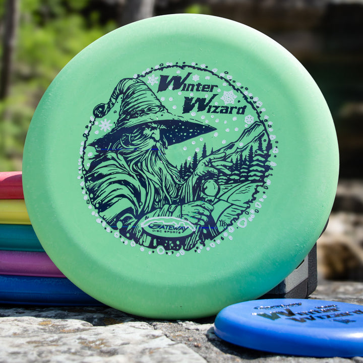 The Winter Wizard Wizard - SSSS + Mini is a green disc golf disc labeled "Winter Wizard" with a wizard illustration, accompanied by a mini—ideal for players seeking max weight and great flight performance.