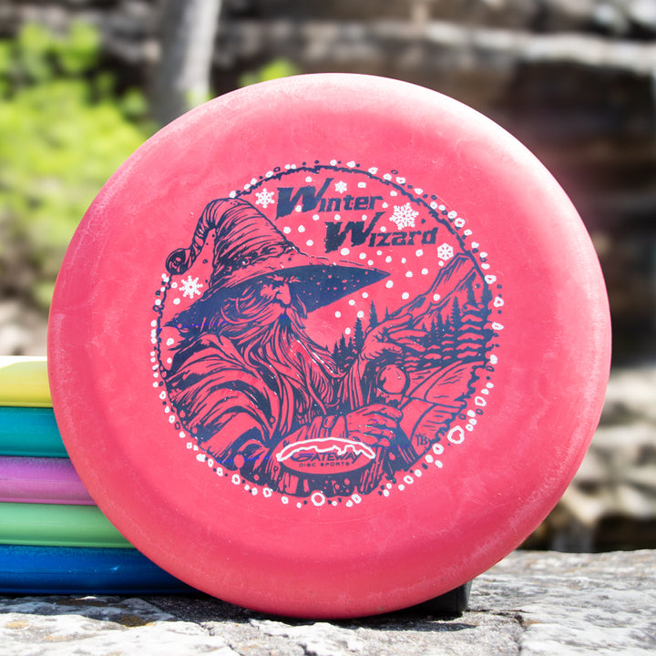 A red Winter Wizard Wizard - SSSS + Mini disc golf disc featuring a hat-wearing wizard with snowflakes, shown atop other discs. Includes max weight and flight numbers for optimal play.