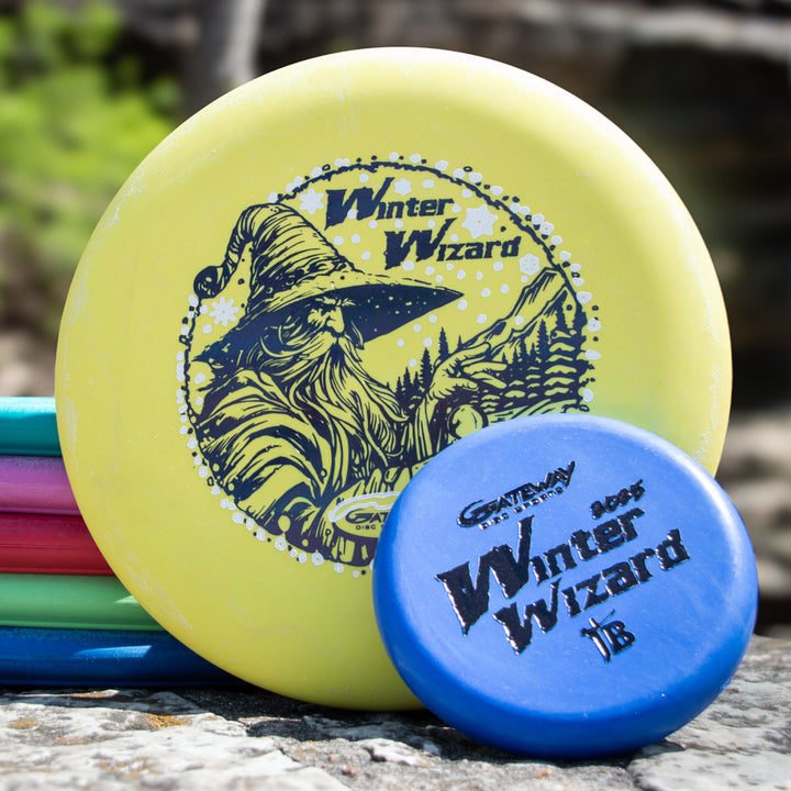 A Winter Wizard Wizard - SSSS + Mini disc set, featuring a yellow disc and blue mini, both labeled with a wizard graphic and flight numbers, rests on a rock with other colorful discs visible in the background.