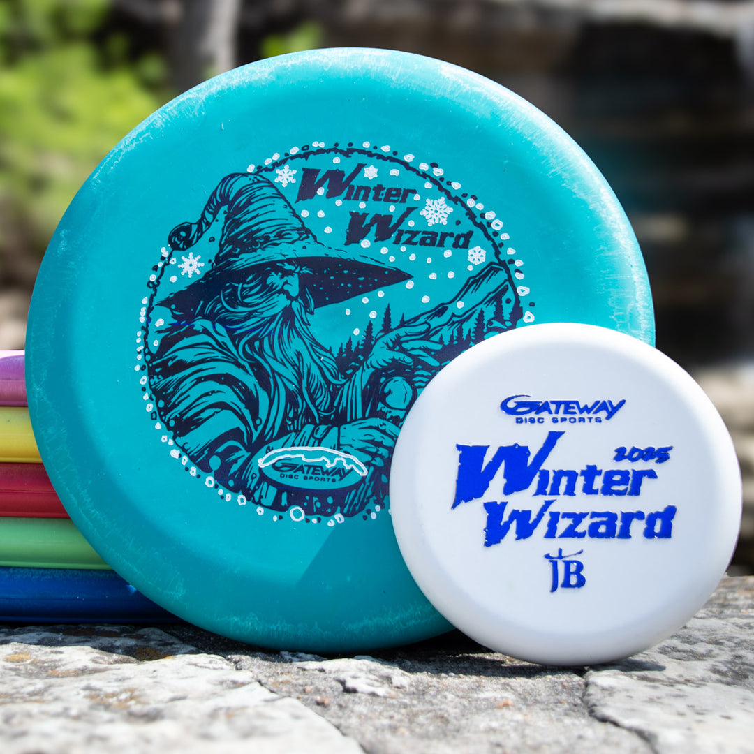Two Max weight Winter Wizard Wizard - SSSS + Mini discs—a turquoise 4S with wizard art and a white with blue text—are stacked outdoors among other colorful discs.