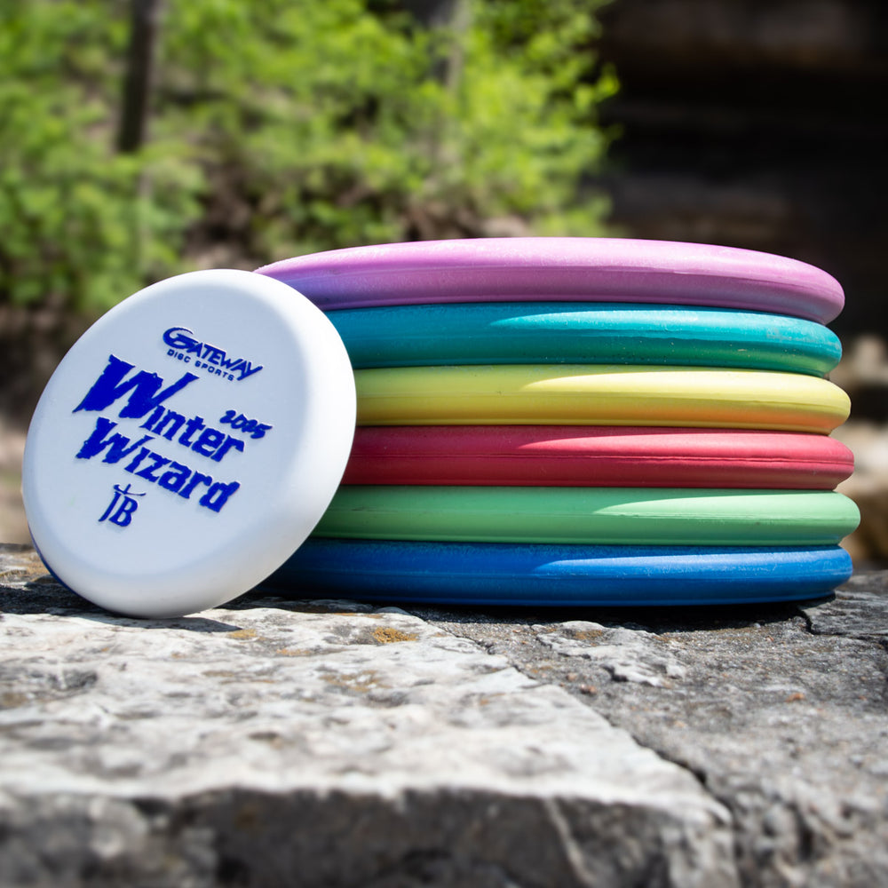 A stack of colorful disc golf discs sits on a rock outdoors, with a white Winter Wizard Wizard - SSSS + Mini at max weight leaning against the stack, ready to reveal its flight numbers.